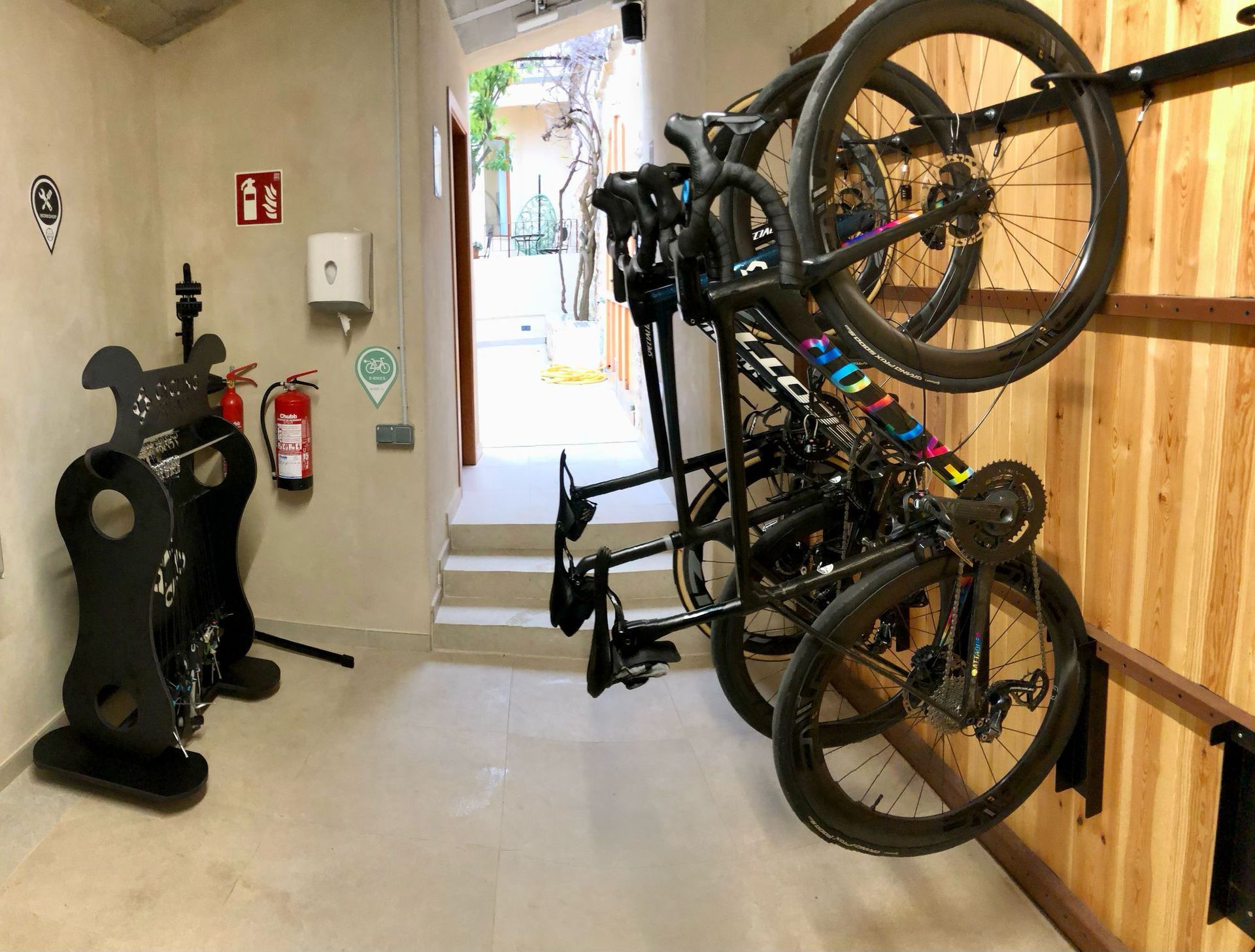 Bicicletas colgadas en una pared de madera, soporte de reparación, extintor y entrada visibles.