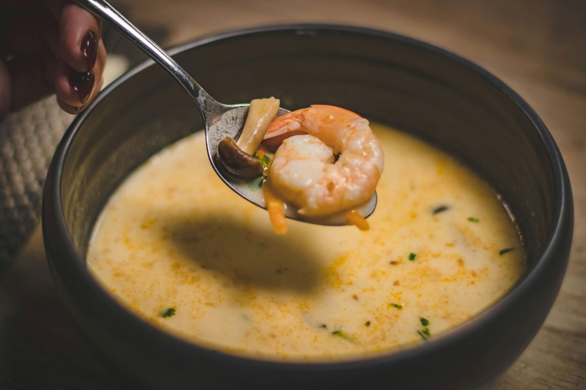 Una persona sostiene una cuchara con un camarón dentro sobre un tazón de sopa.